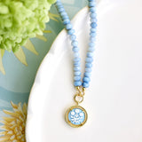 A light blue beaded necklace with a round gold pendant featuring a blue and white floral design, styled on a white ceramic dish with green flowers in the background.
