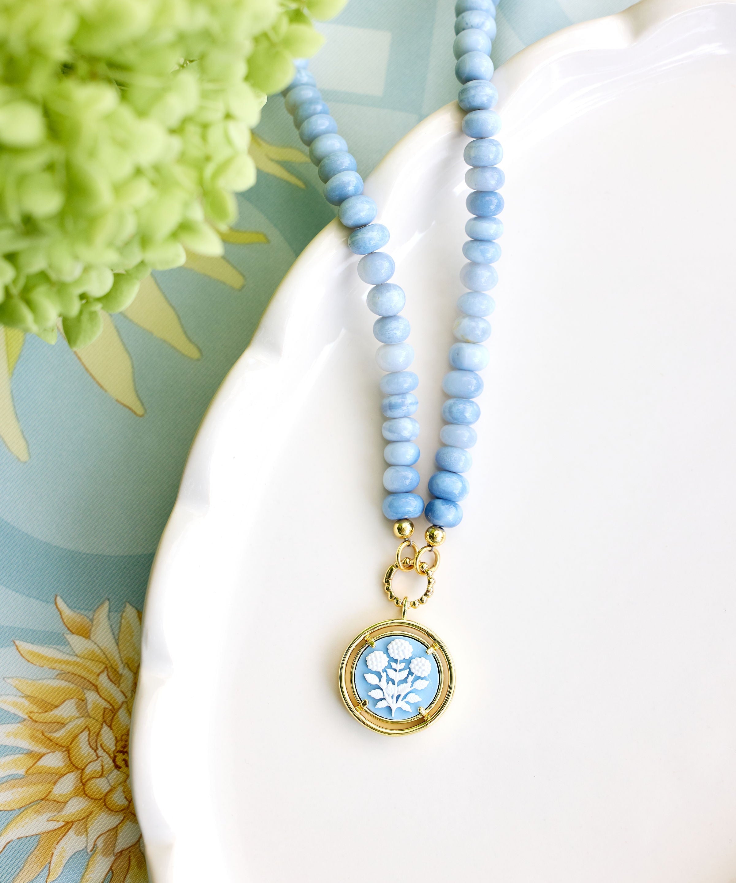 A light blue beaded necklace with a round gold pendant featuring a blue and white floral design, styled on a white ceramic dish with green flowers in the background.