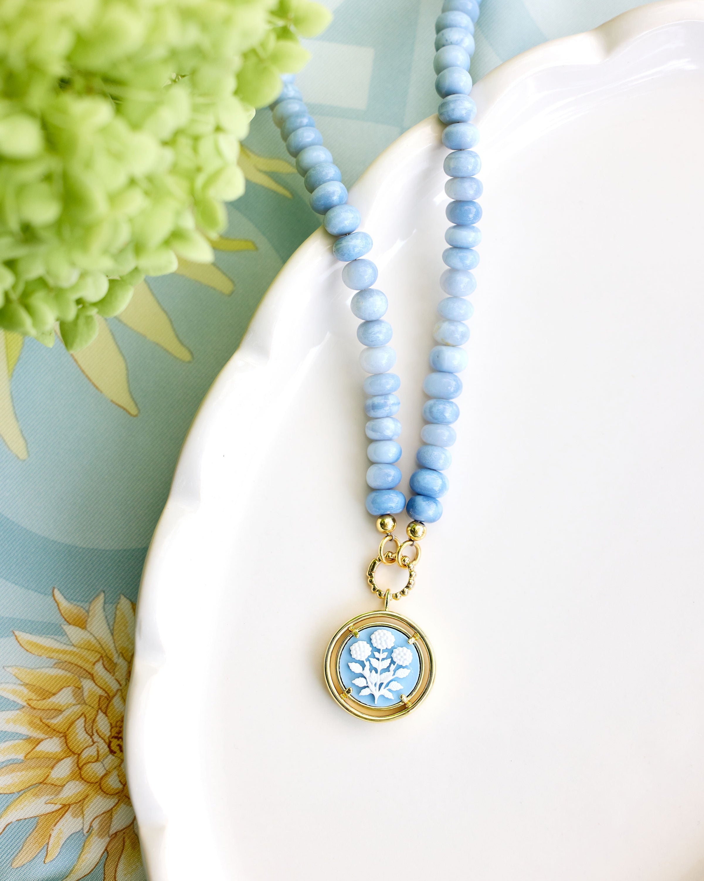 A light blue beaded necklace with a round gold pendant featuring a blue and white floral design, styled on a white ceramic dish with green flowers in the background.