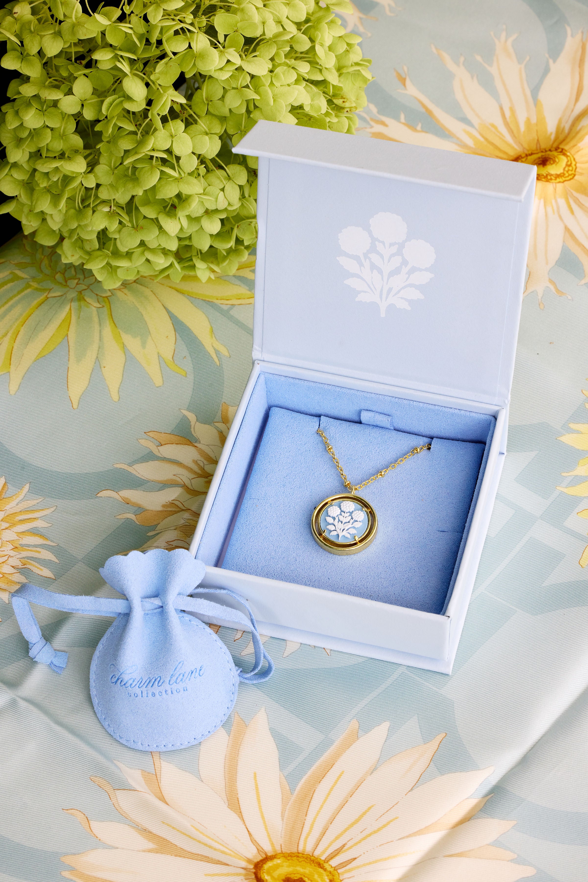 A gold cameo necklace displayed in an open blue jewelry box with a light blue velvet interior, alongside a small light blue drawstring pouch. The scene is styled with green and yellow flowers on a patterned surface.