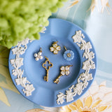 A styled flat-lay of several small jewelry pieces arranged on a light blue decorative plate with white floral relief. The jewelry includes gold-tone key-shaped and flower-shaped pieces with clear stones, pearl-like white bead clusters, and a light blue flower charm.