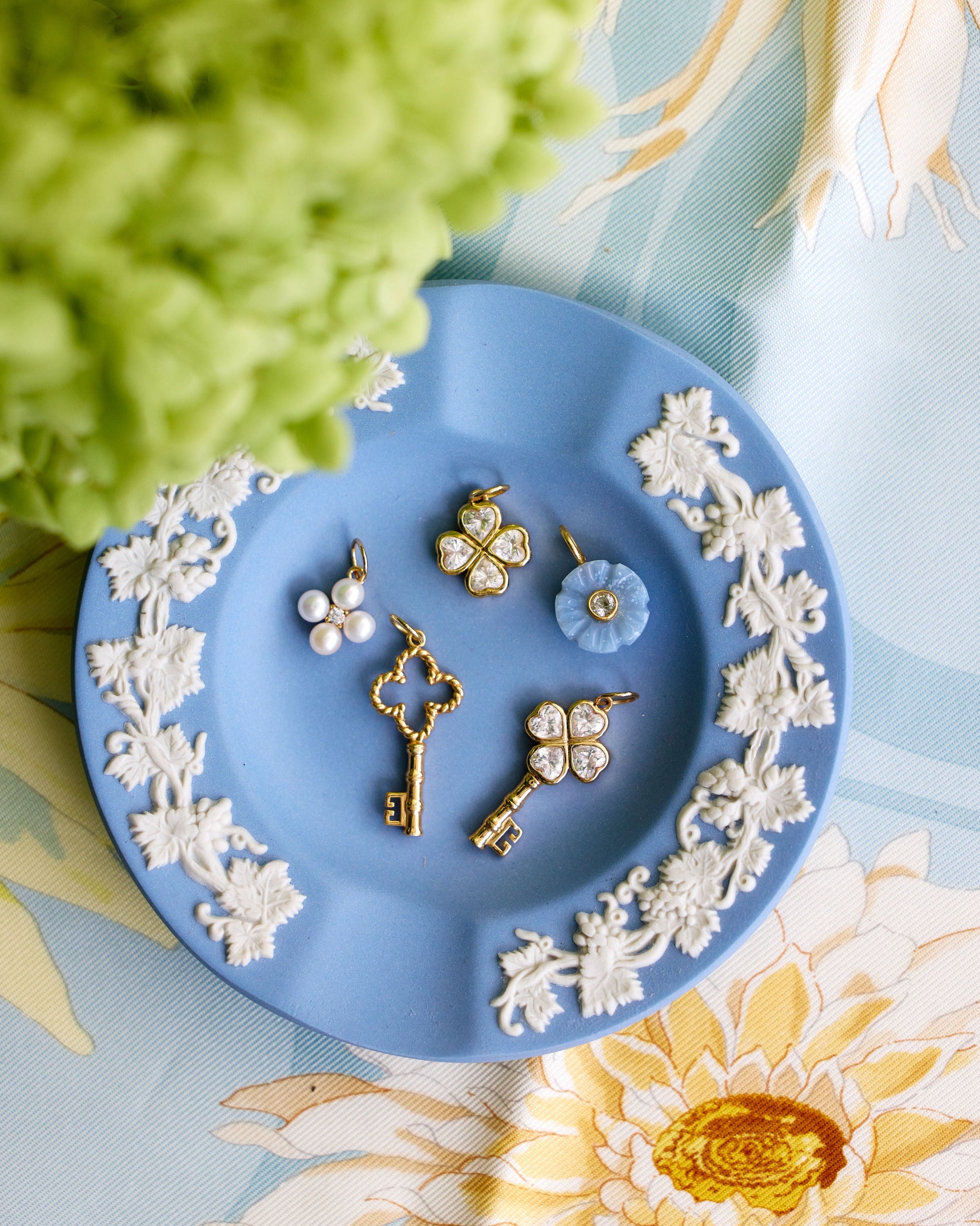 A styled flat-lay of several small jewelry pieces arranged on a light blue decorative plate with white floral relief. The jewelry includes gold-tone key-shaped and flower-shaped pieces with clear stones, pearl-like white bead clusters, and a light blue flower charm.
