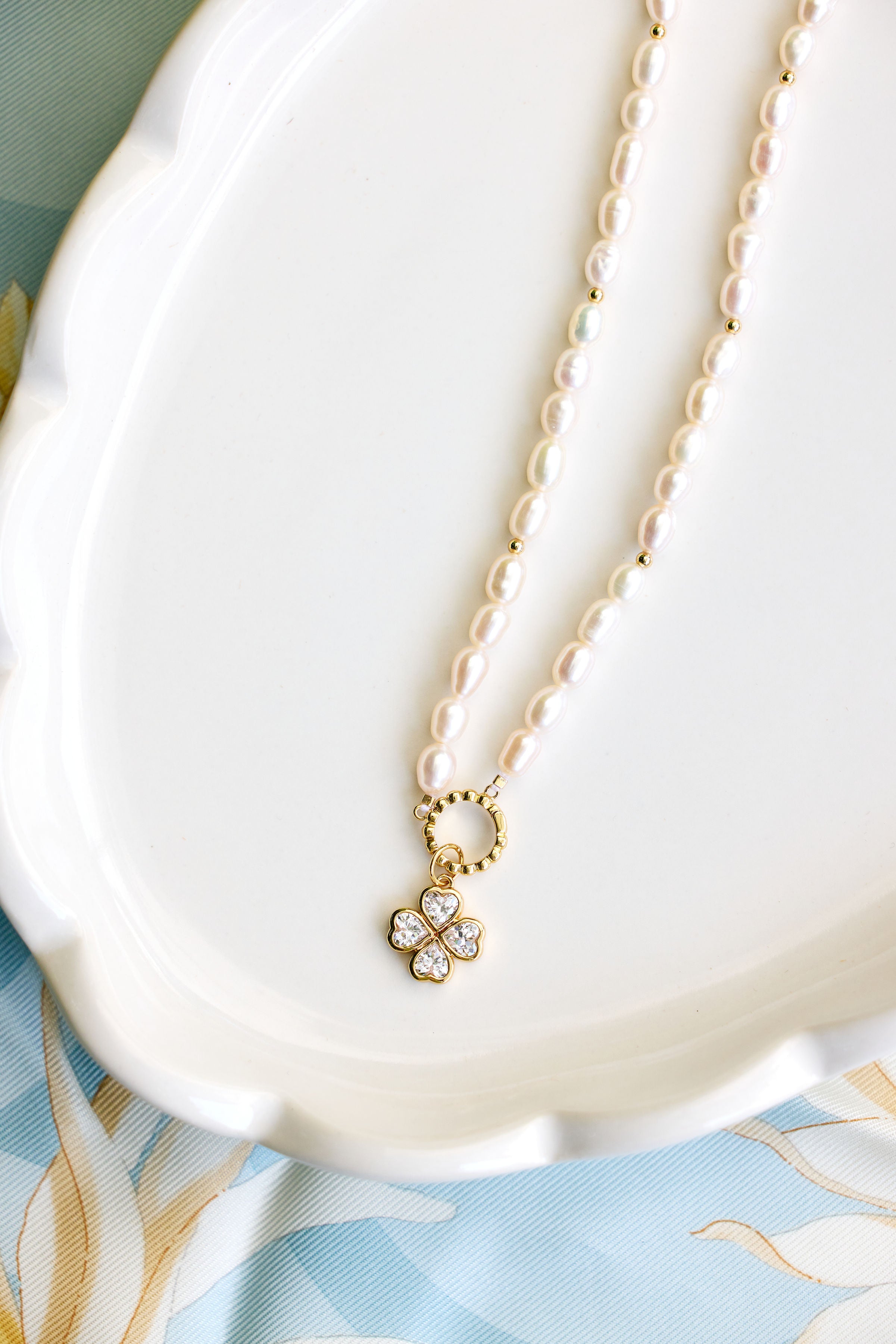 A delicate necklace with small white pearls and a gold four-leaf clover pendant set with clear crystals, resting on a white scalloped dish.