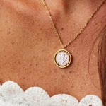 A gold chain necklace with a circular pendant featuring a carved white floral, worn on a person's neck above a white lace top.