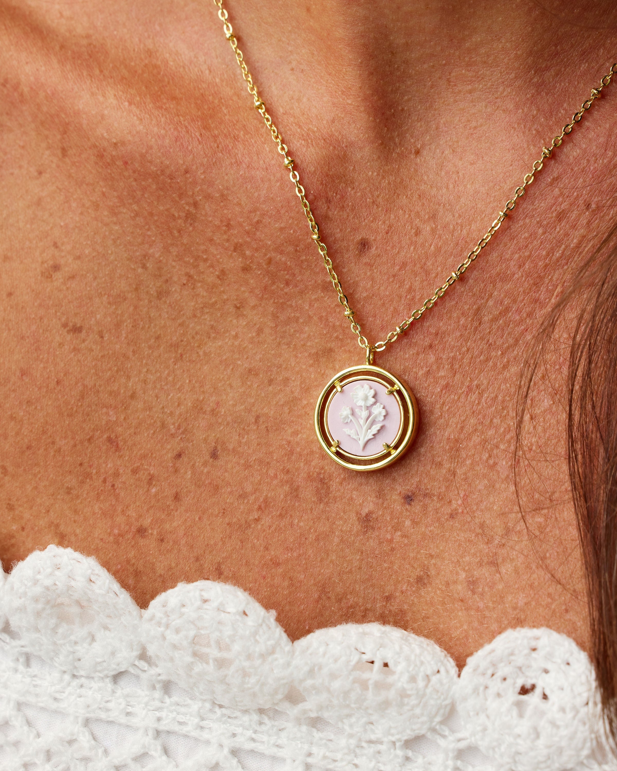A gold chain necklace with a circular pendant featuring a carved white floral, worn on a person's neck above a white lace top.