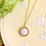 A gold pendant necklace with a circular frame and a pale pink floral cameo pendant, displayed on a yellow decorative tray with white floral embossing and soft green foliage in the corner.