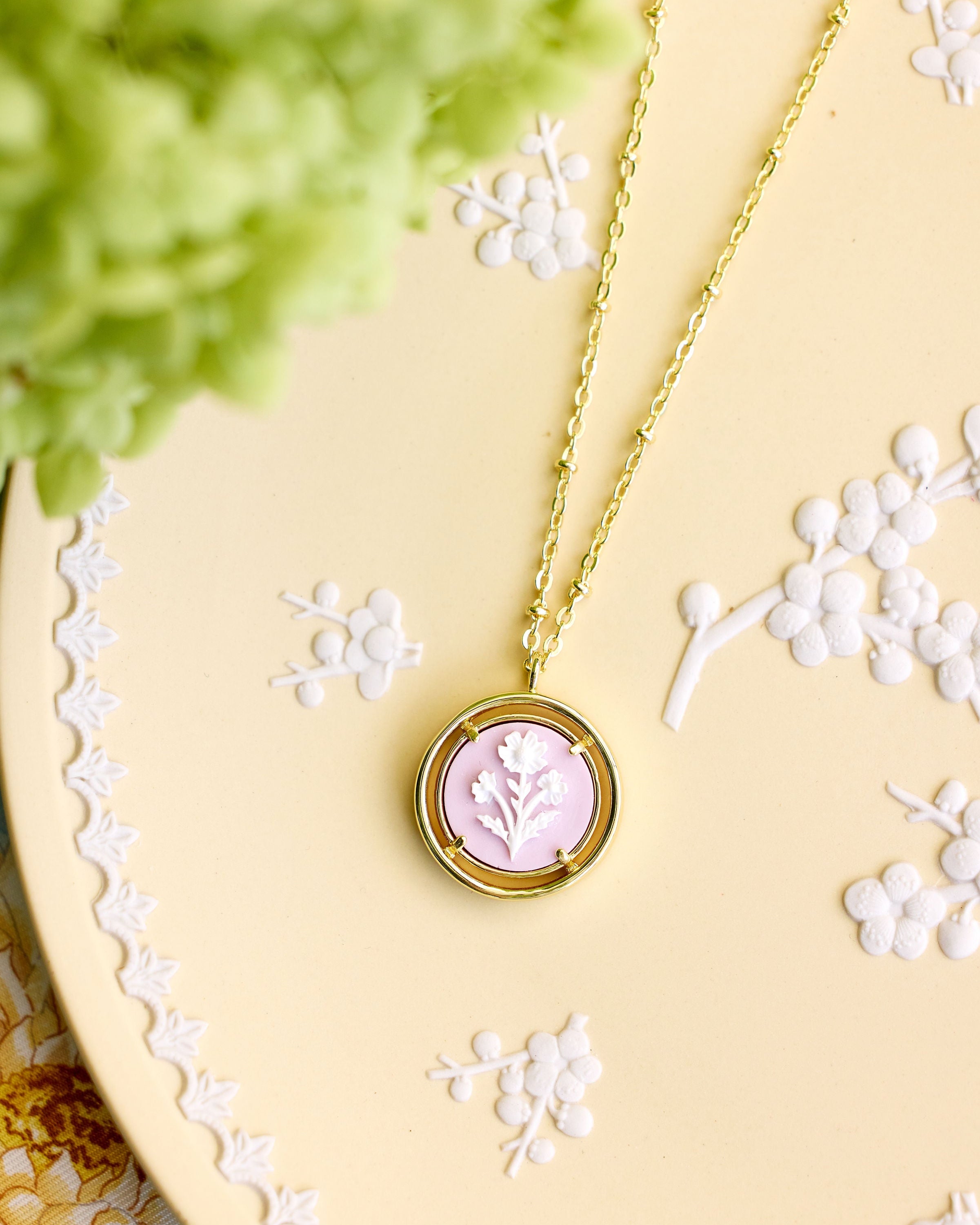 A gold pendant necklace with a circular frame and a pale pink floral cameo pendant, displayed on a yellow decorative tray with white floral embossing and soft green foliage in the corner.