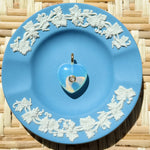 Blue decorative plate with white floral patterns on a woven surface displaying a heart charm