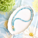 light aqua beaded necklace with a green heart pendant styled on a white dish on a floral background
