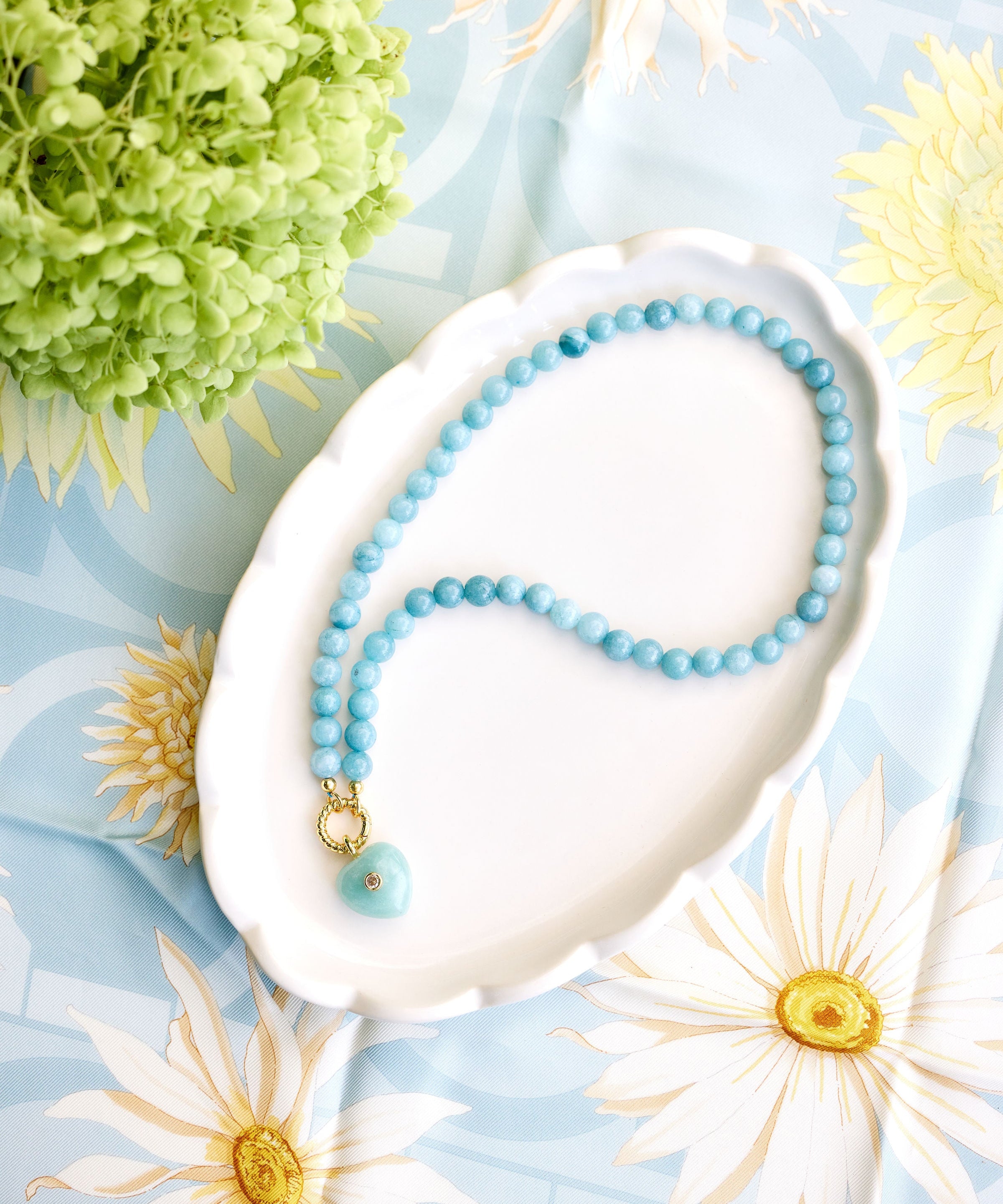 light aqua beaded necklace with a green heart pendant styled on a white dish on a floral background