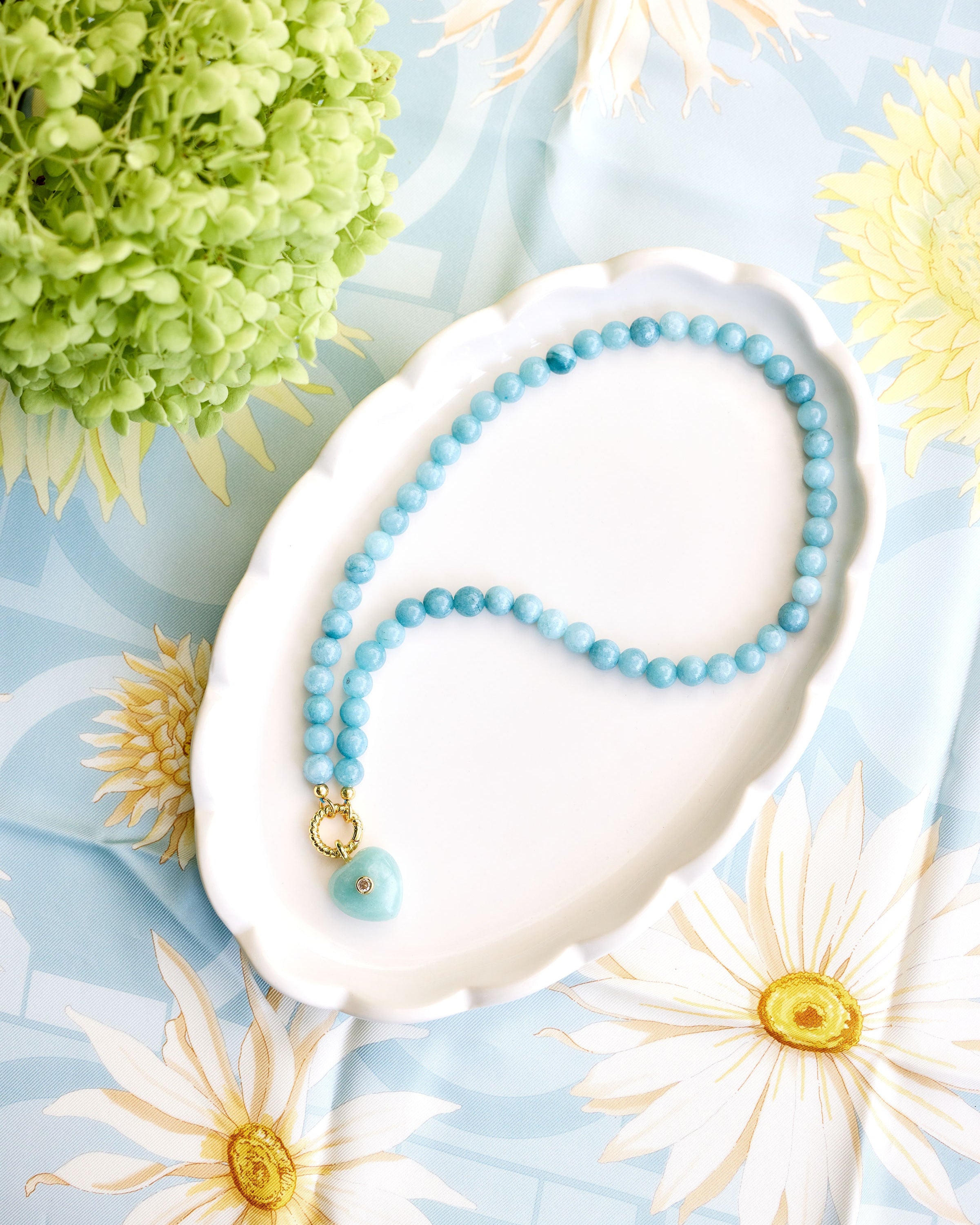 light aqua beaded necklace with a green heart pendant styled on a white dish on a floral background