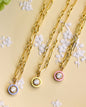 Three gold chain necklaces with small oval pendant charms in blue, yellow, and pink, arranged on a beige decorative plate with white floral patterns and green hydrangea flowers.