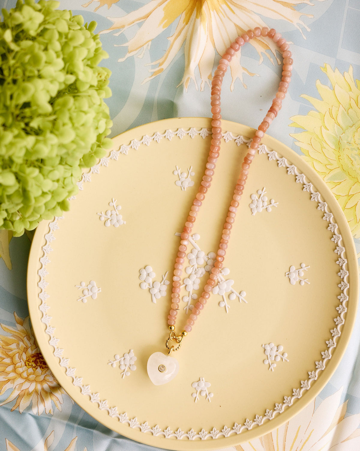 A peach-pink beaded necklace with a white heart-shaped pendant and gold clasp, displayed on a round cream-colored plate with floral patterns, surrounded by green and yellow flowers on a blue patterned fabric background.