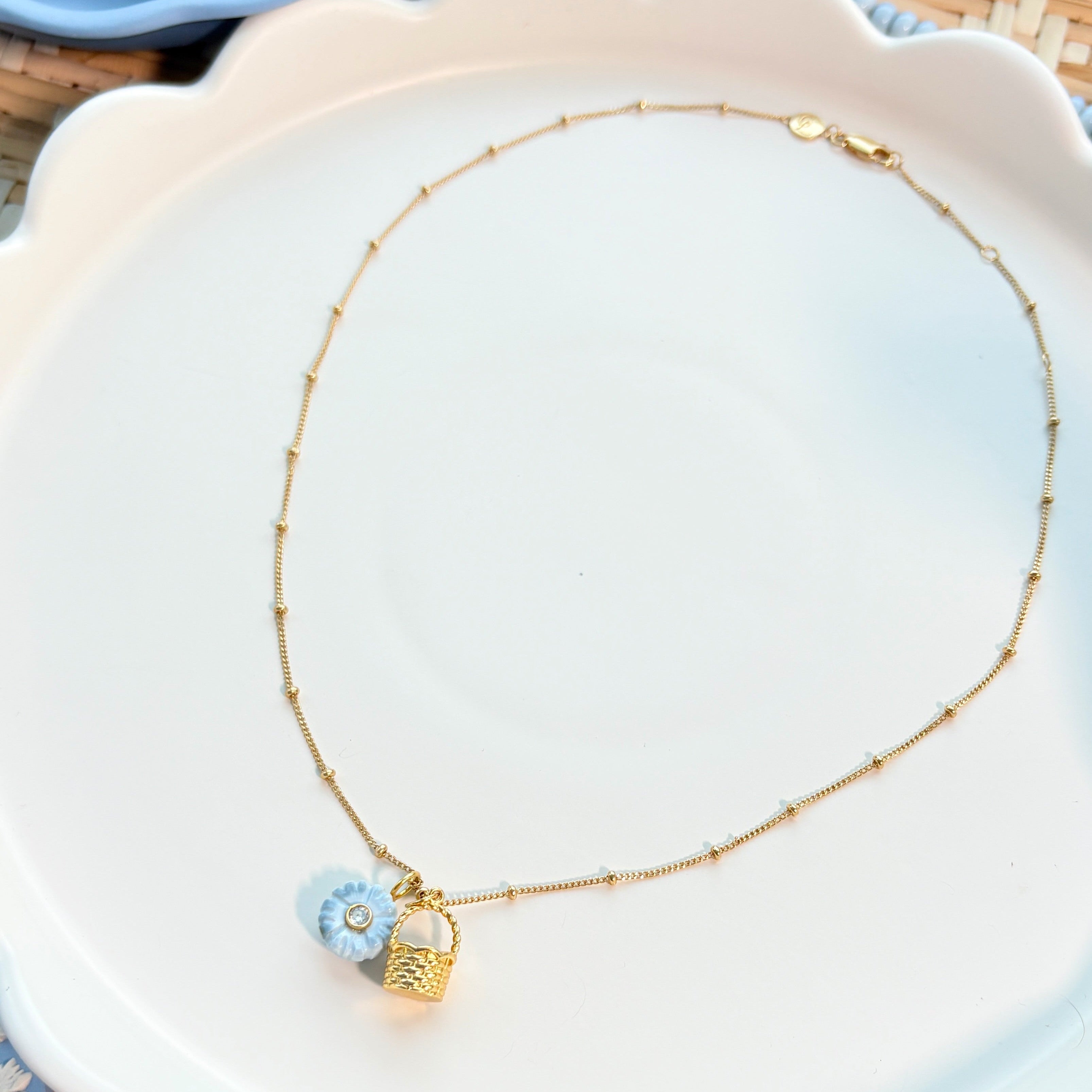 Necklace with a gold basket charm and blue flower charm on a white scalloped plate
