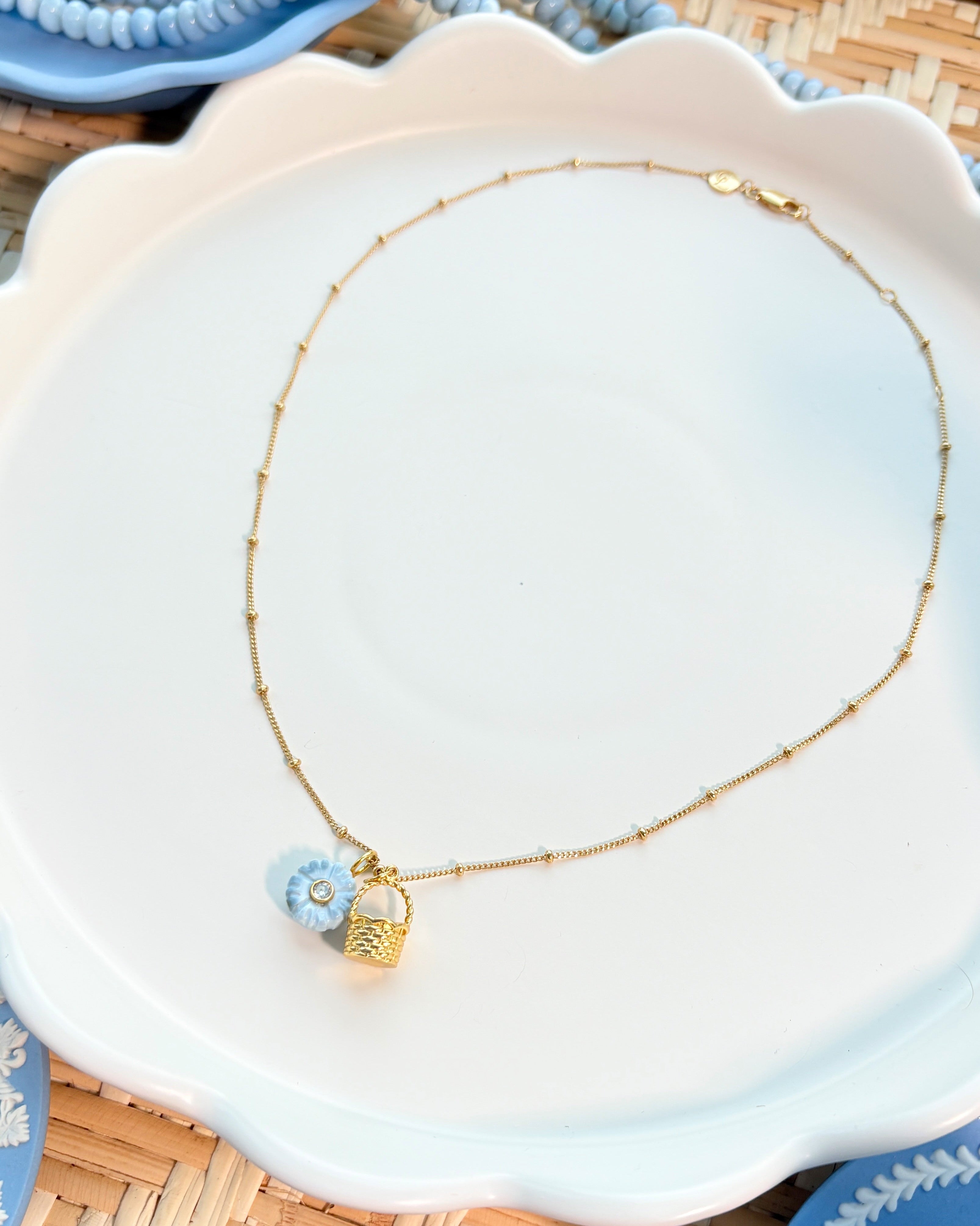 Necklace with a gold basket charm and blue flower charm on a white scalloped plate