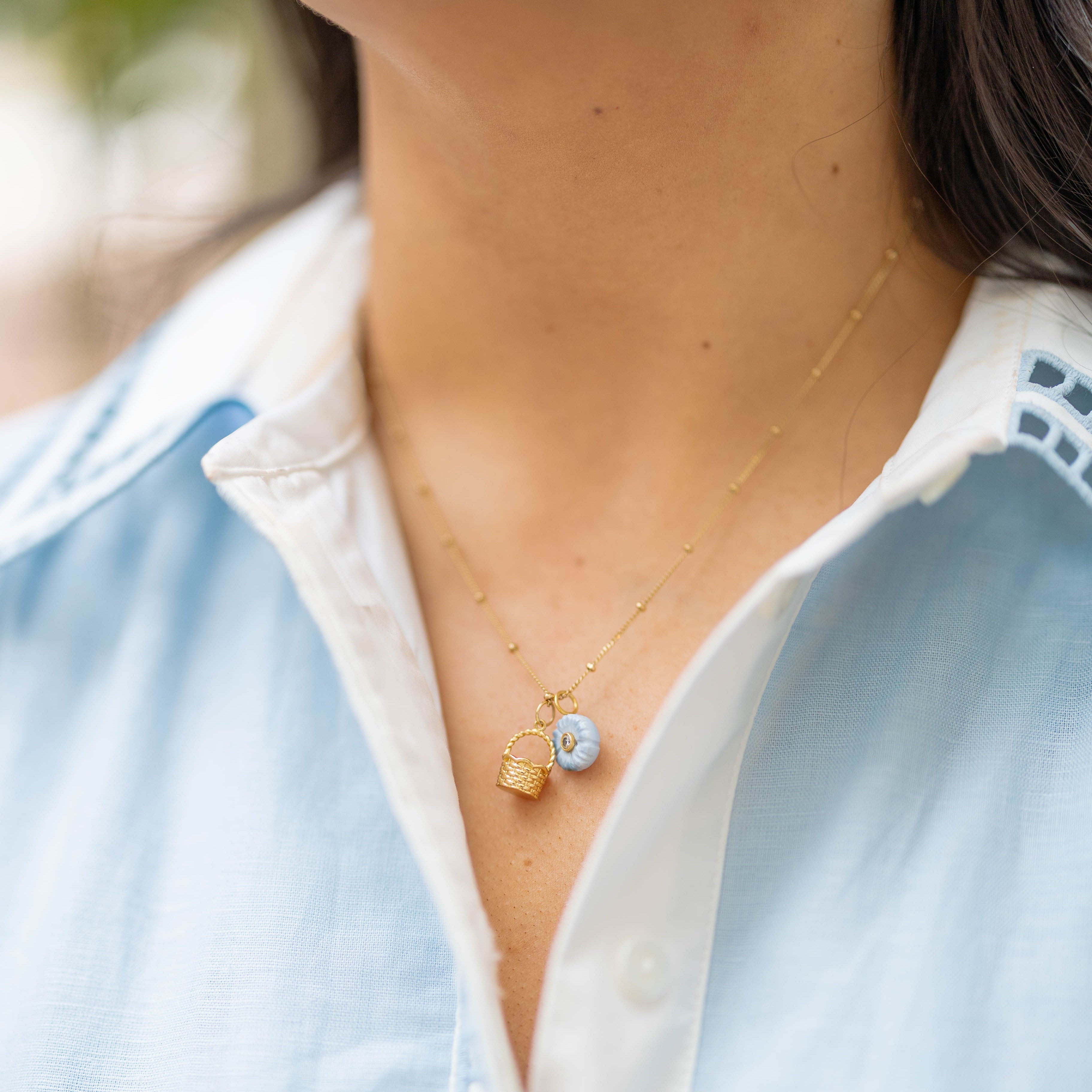 Necklace with a gold basket charm and blue flower charm on a person wearing a blue collared shirt