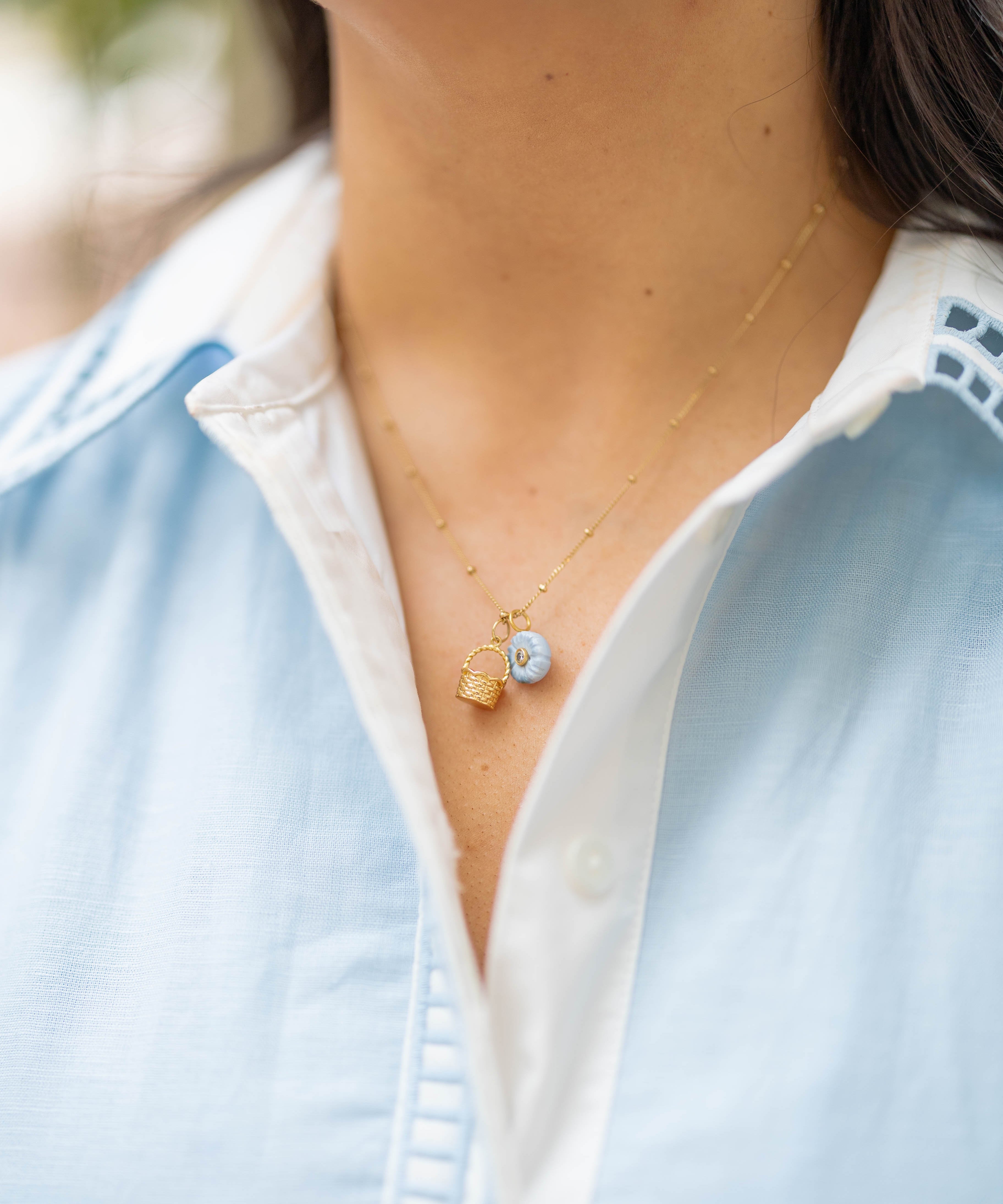 Necklace with a gold basket charm and blue flower charm on a person wearing a blue collared shirt