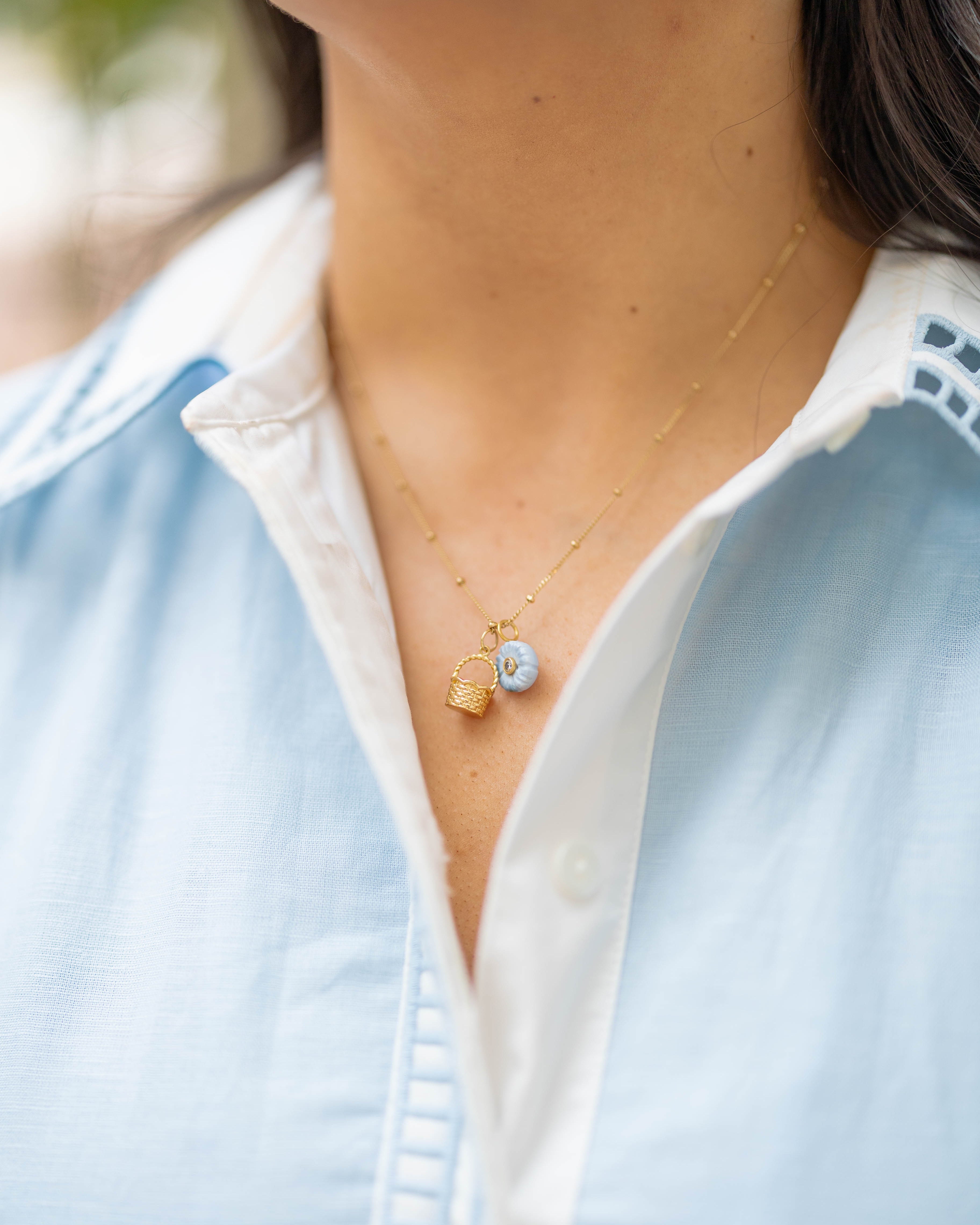 Necklace with a gold basket charm and blue flower charm on a person wearing a blue collared shirt