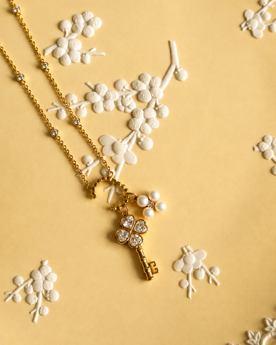 A gold key-shaped pendant and pearl clover necklace with small clear stones, displayed on a warm yellow surface decorated with white floral relief details.