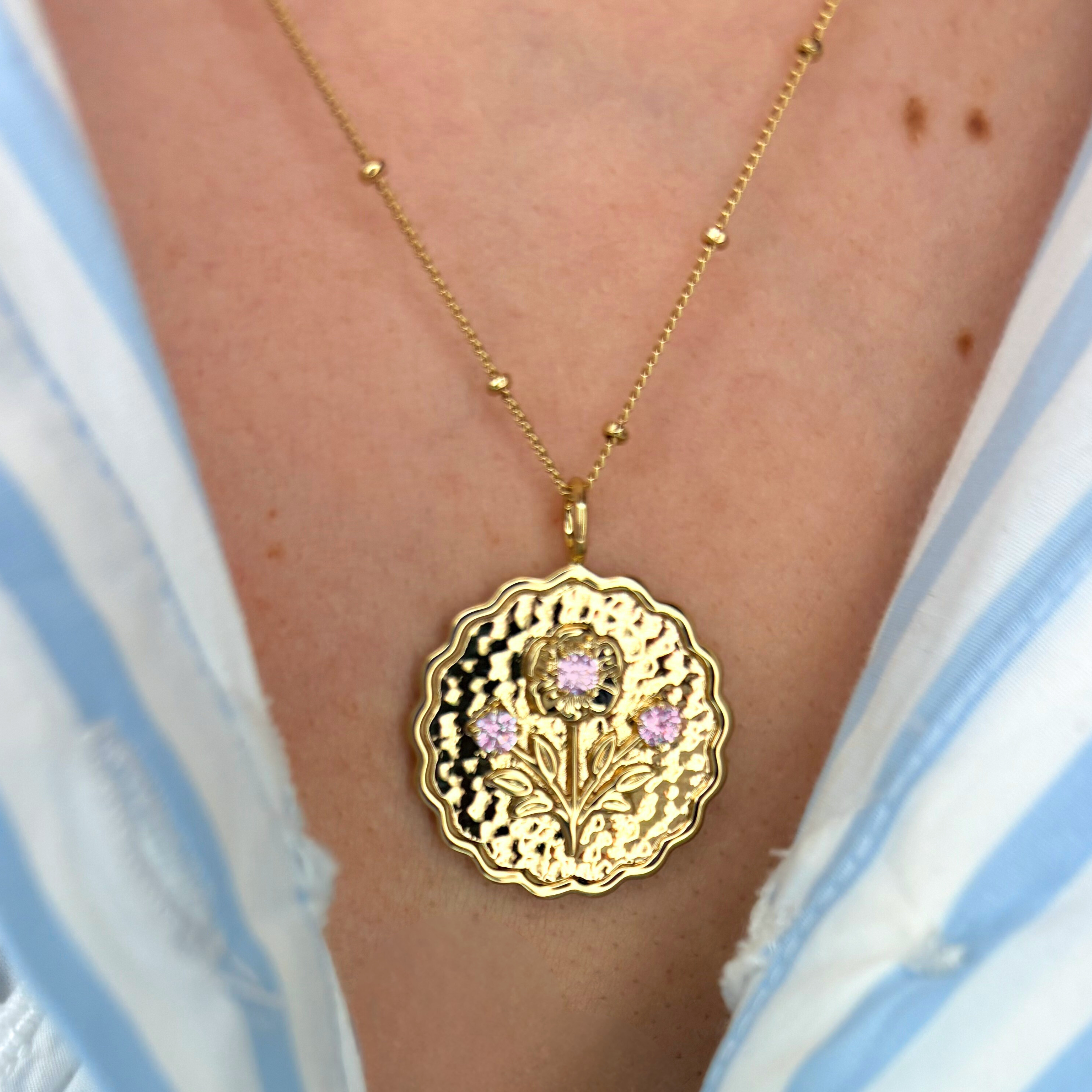 Gold necklace with a floral pendant featuring pink gemstones on a person wearing a striped garment.