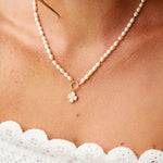 A close-up of a woman wearing a delicate pearl necklace with a small gold flower charm, paired with a white lace top.