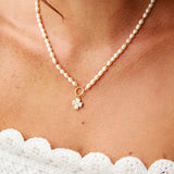 A close-up of a woman wearing a delicate pearl necklace with a small gold flower charm, paired with a white lace top.