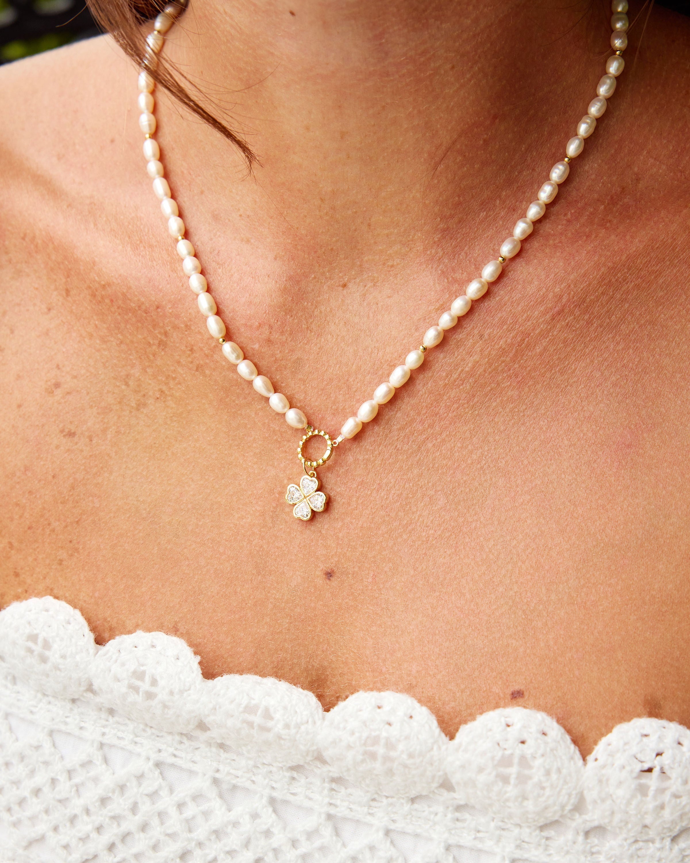 A close-up of a woman wearing a delicate pearl necklace with a small gold flower charm, paired with a white lace top.