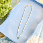 A white pearl necklace with a gold clasp and a small gold ring pendant, displayed on a light blue rectangular tray with floral props.