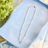A white pearl necklace with a gold clasp and a small gold ring pendant, displayed on a light blue rectangular tray with floral props.