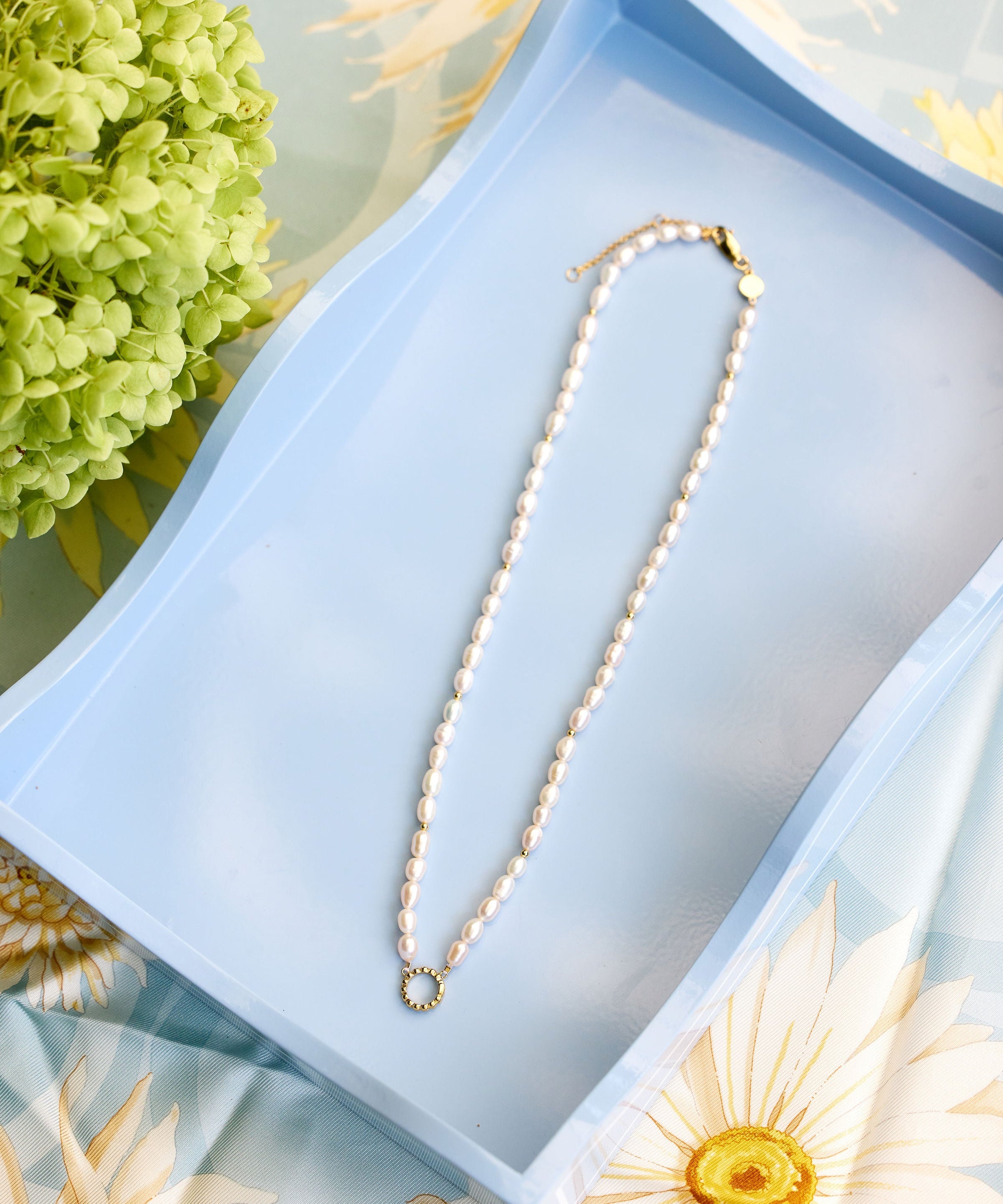 A white pearl necklace with a gold clasp and a small gold ring pendant, displayed on a light blue rectangular tray with floral props.