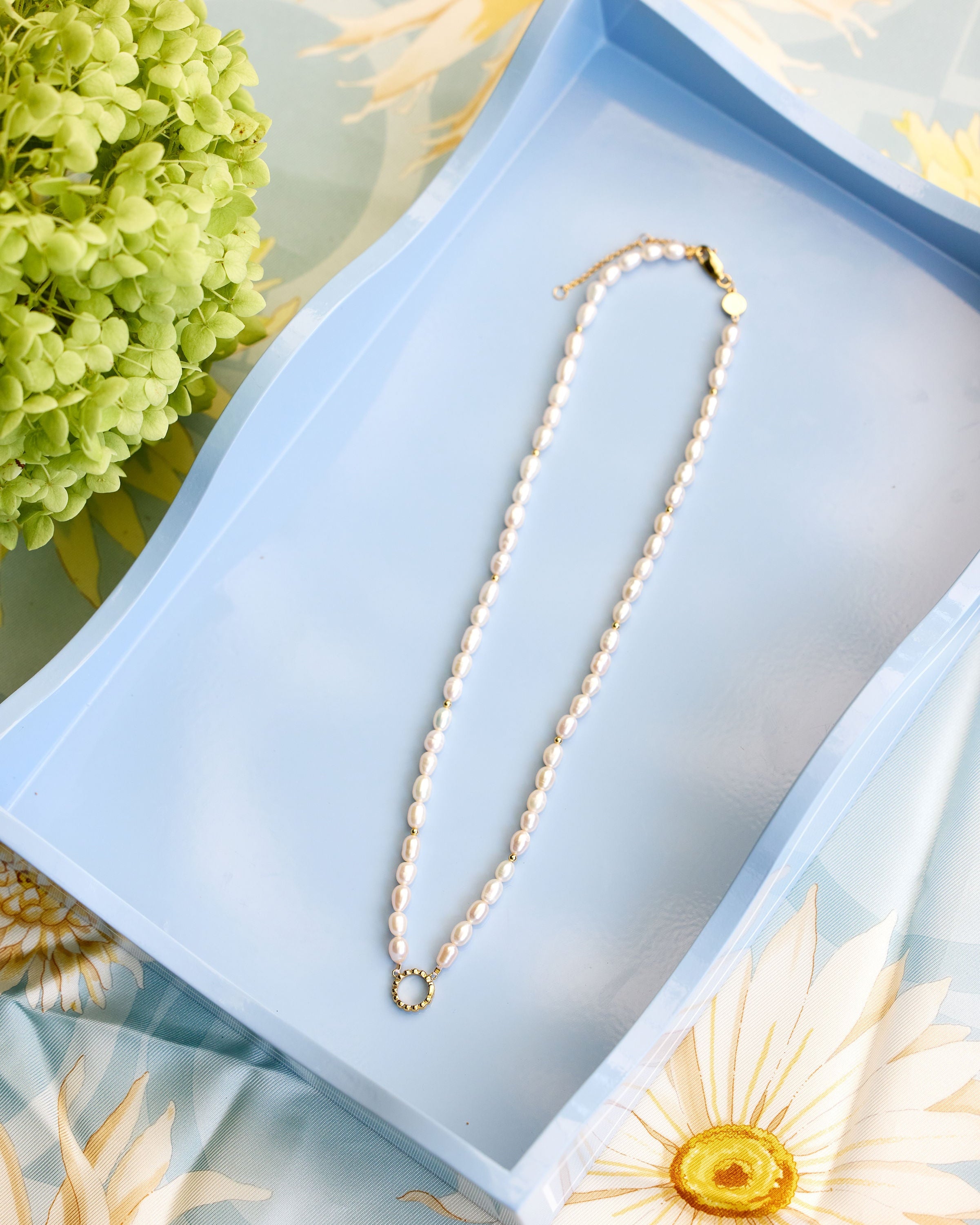 A white pearl necklace with a gold clasp and a small gold ring pendant, displayed on a light blue rectangular tray with floral props.