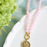 A pink beaded necklace with a textured gold coin pendant, draped over a white ceramic dish with a green hydrangea in the background.