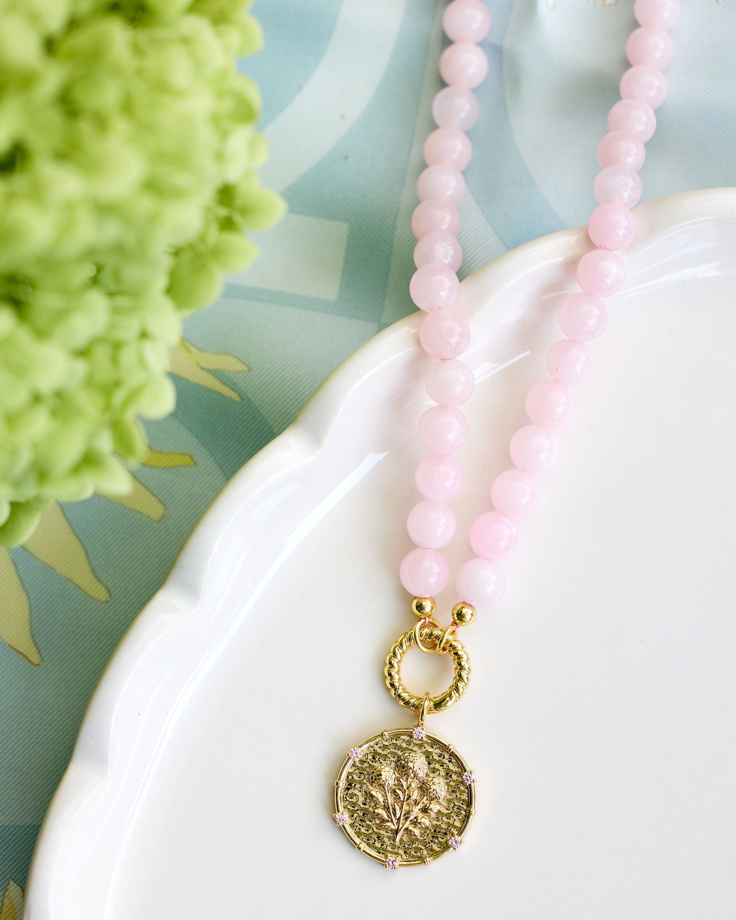 A pink beaded necklace with a textured gold coin pendant, draped over a white ceramic dish with a green hydrangea in the background.