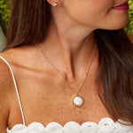 A woman wearing a delicate gold necklace with a round pink cameo pendant and a matching stud earring, shown in a close-up profile view.