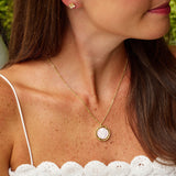 A woman wearing a delicate gold necklace with a round pink cameo pendant and a matching stud earring, shown in a close-up profile view.