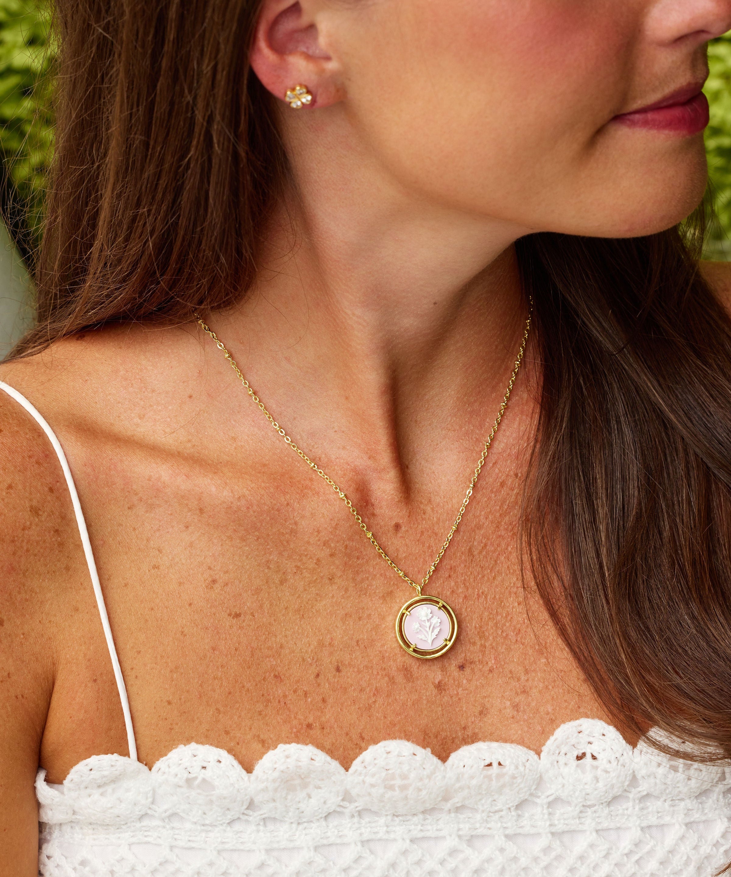 A woman wearing a delicate gold necklace with a round pink cameo pendant and a matching stud earring, shown in a close-up profile view.