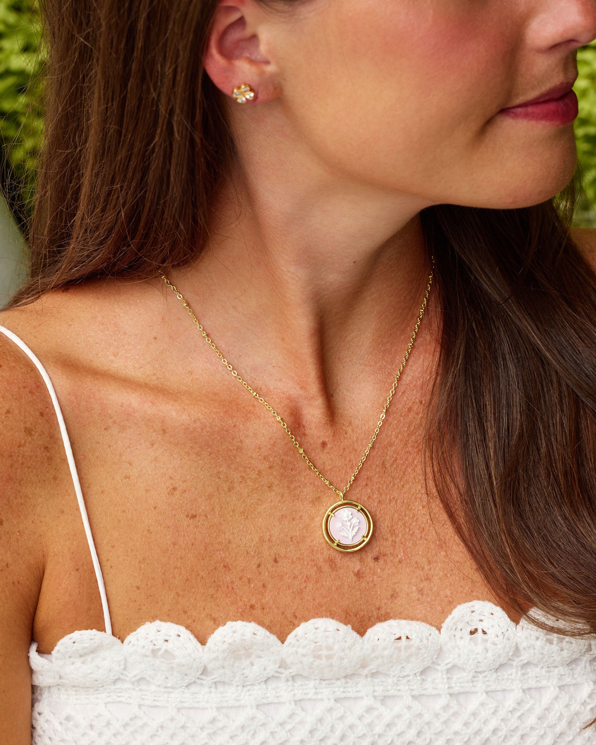 A woman wearing a delicate gold necklace with a round pink cameo pendant and a matching stud earring, shown in a close-up profile view.