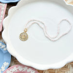 gold scalloped charm with pink gemstones on a pink beaded chain displayed on a white tray