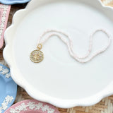 gold scalloped charm with pink gemstones on a pink beaded chain displayed on a white tray