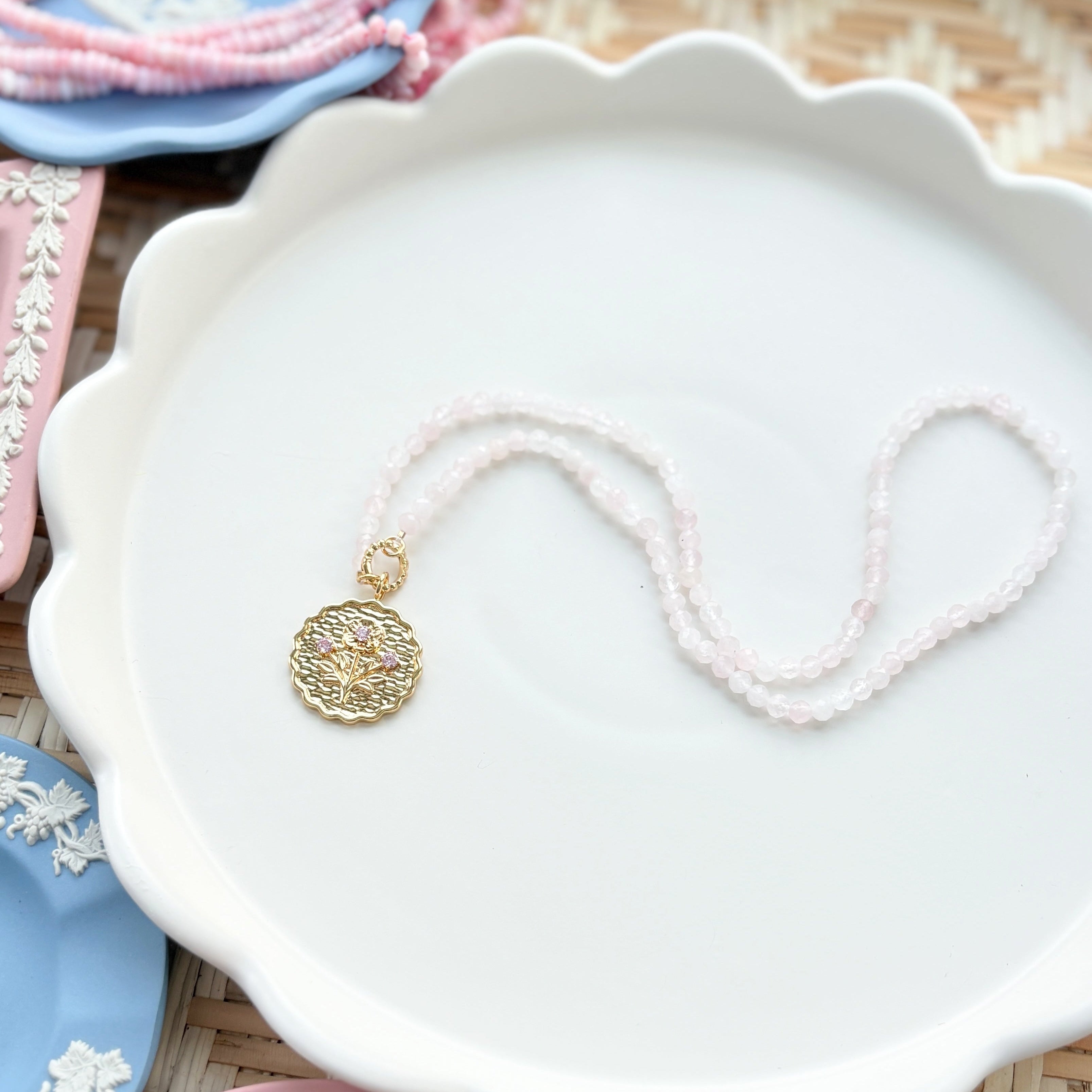 gold scalloped charm with pink gemstones on a pink beaded chain displayed on a white tray