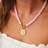 A close-up of a model wearing a pink beaded necklace with a gold-tone circular pendant and a small gold ring clasp. The model is wearing a white textured top.