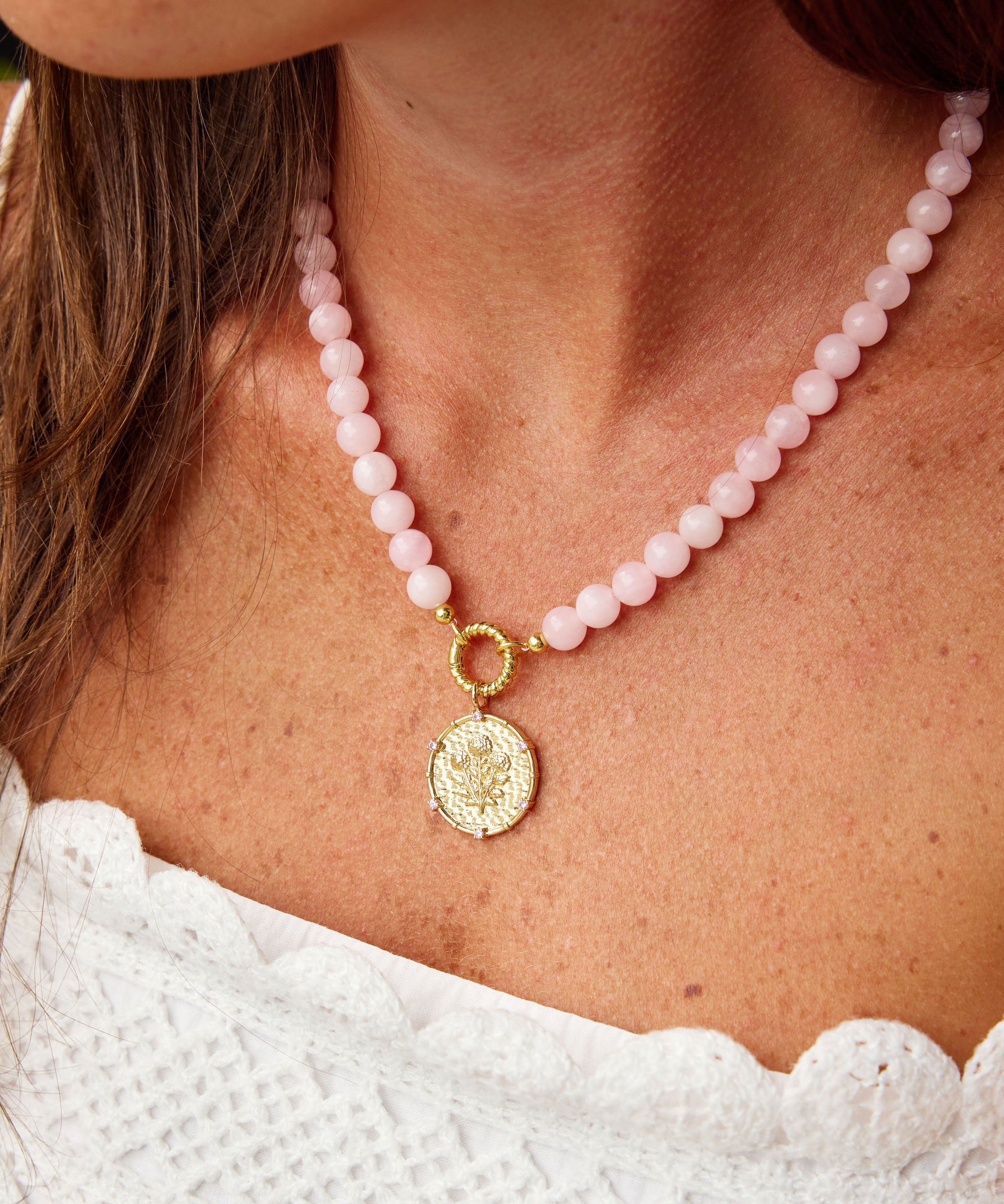 A close-up of a model wearing a pink beaded necklace with a gold-tone circular pendant and a small gold ring clasp. The model is wearing a white textured top.
