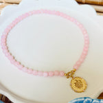 Pink beaded necklace with a golden coin pendant on a white scalloped tray