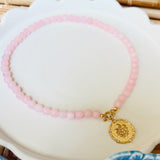 Pink beaded necklace with a golden coin pendant on a white scalloped tray