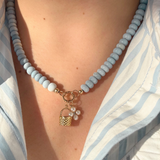 Necklace with light blue beads and a gold basket charm and pearl flower charm worn by a person in a blue and white striped shirt