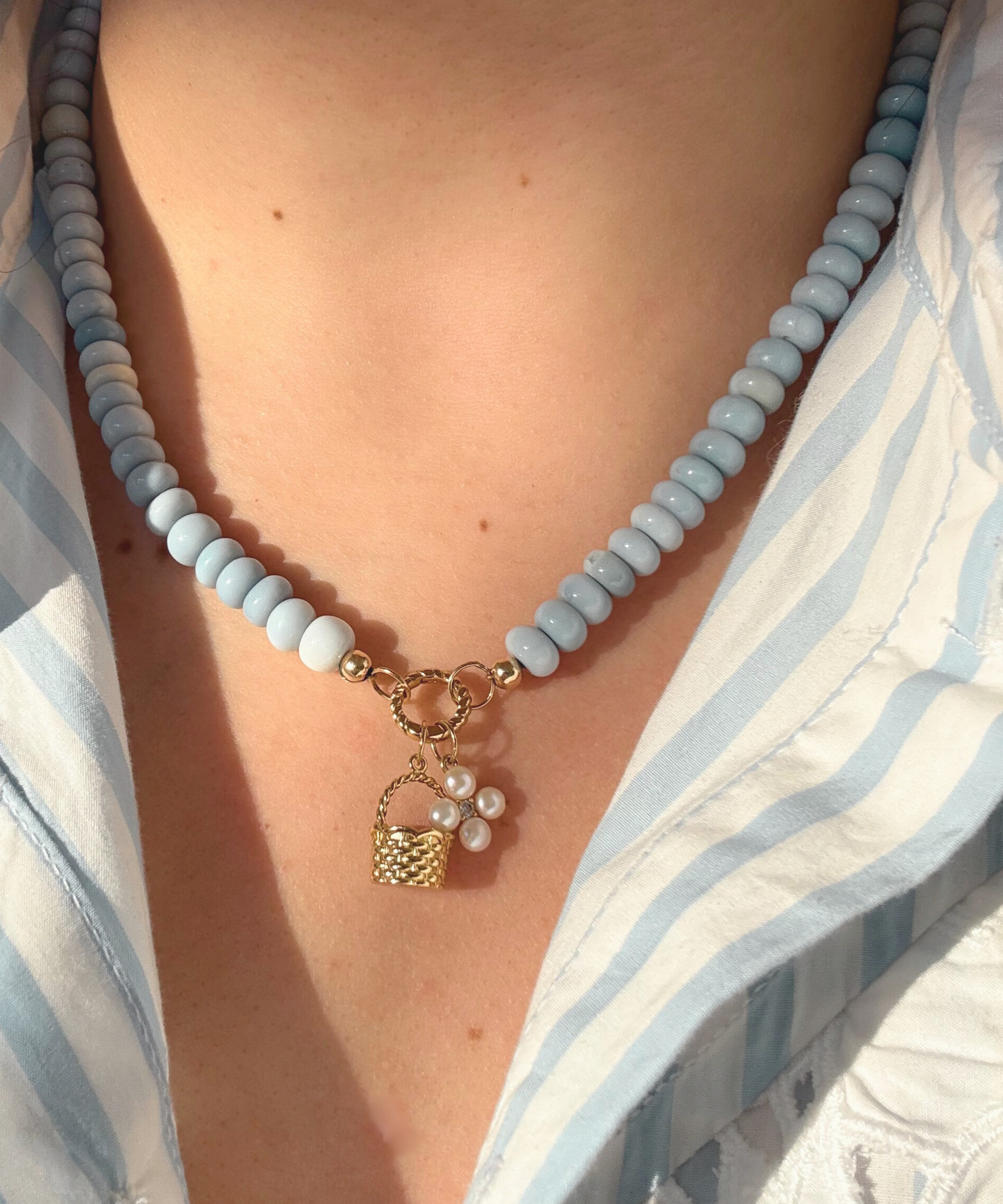 Necklace with light blue beads and a gold basket charm and pearl flower charm worn by a person in a blue and white striped shirt