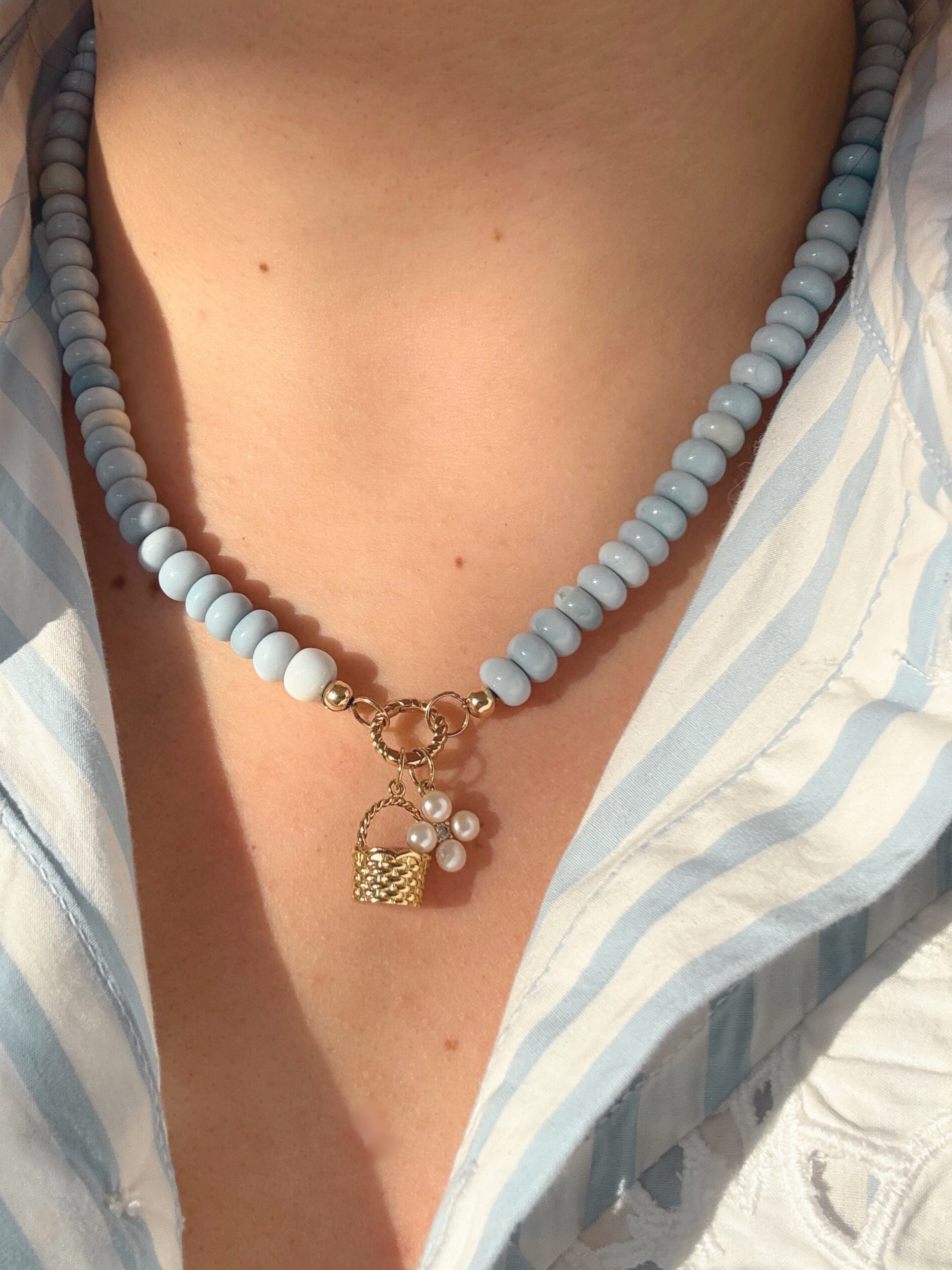 Necklace with light blue beads and a gold basket charm and pearl flower charm worn by a person in a blue and white striped shirt