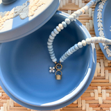 Blue jewelry box with a beaded necklace  with a pearl flower and gold basket charm on a woven surface