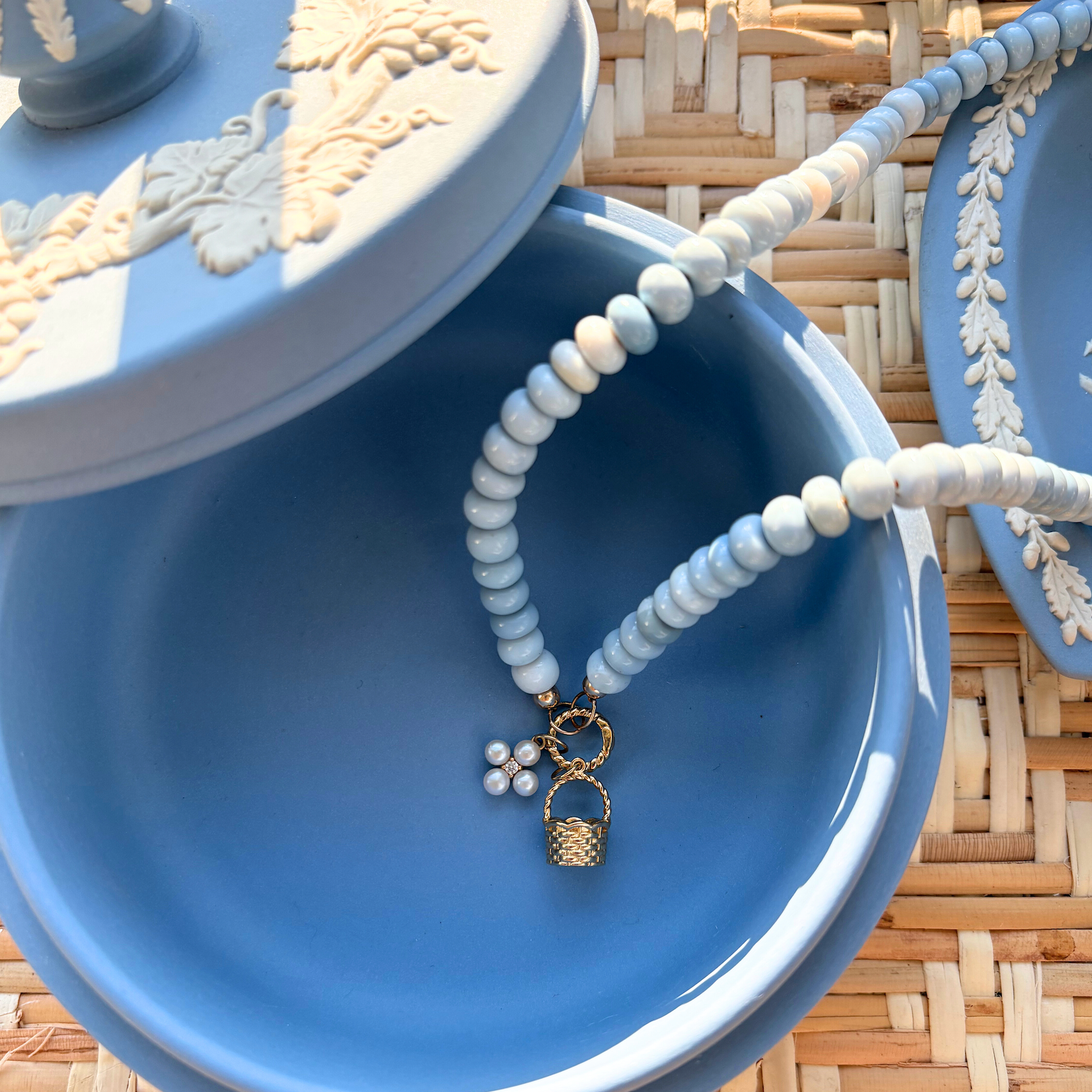 Blue jewelry box with a beaded necklace  with a pearl flower and gold basket charm on a woven surface