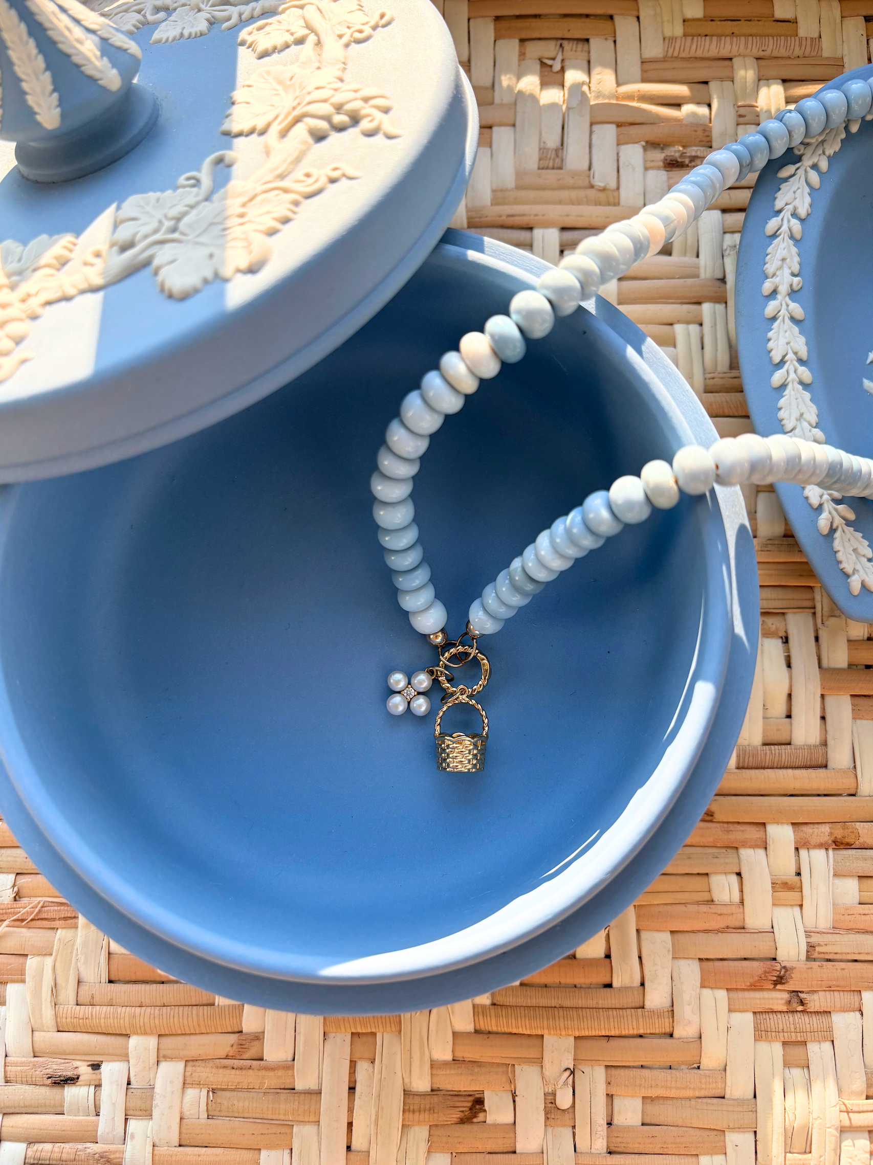 Blue jewelry box with a beaded necklace  with a pearl flower and gold basket charm on a woven surface